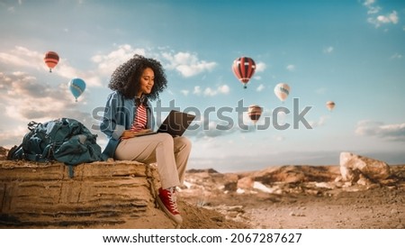 Excited Beautiful Young Multiethnic Female Traveller with Afro Hairstyle Using Laptop Computer on Top of Rocky Canyon Valley. Hot Air Balloons Fly in Mountain National Park. Hiking in Nature.