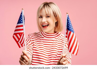 Excited, beautiful young woman with blonde hair holding American flags looking at camera. Attractive female supporter standing isolated on pink background. American independence day concept - Powered by Shutterstock