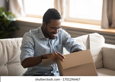 Excited biracial young man sit on couch at home unpack cardboard box with Internet order, happy African American male open carton package at home, shopping on web, good delivery service concept