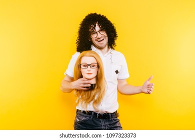 Excited bizarre strange odd foolish guy have fun with plastic mannequin.  Weird relationship. Happy adult man in black curly wig isolated on yellow background. Oddball rebel with hairy girl head