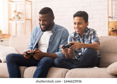 Excited Black Boy Playing Video Games With His Dad, Sitting Together On Couch At Home.