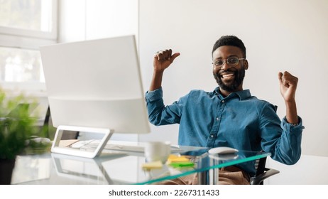 Excited black businessman gesturing yes in front computer  celebrating success  sitting at workplace in office  panorama  Male entrepreneur making great deal  signing online contract