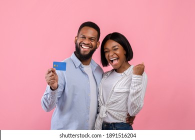 Excited black couple with credit card making YES gesture, promoting contactless shopping over pink studio background. Affectionate young lovers buying gifts for Valentine's Day online, using bank card