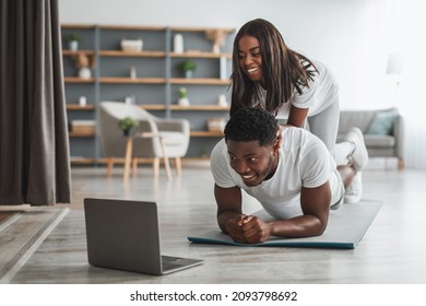 Excited Black Couple Having Fun During Home Workout Looking At Laptop Screen Watching Virtual Tutorial, Boyfriend Doing Plank Exercise With Girlfriend Lying On Him On His Back In Living Room