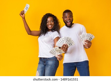 Excited Black Couple Holding Dollars In Both Hands, Yellow Studio Background