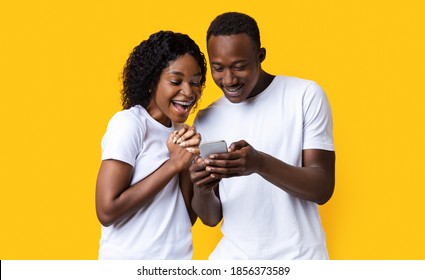 Excited Black Couple Looking At Mobile Phone Screen, Using New Cool Application, Yellow Studio Background. Joyful African American Man And Woman Using Smartphone Together, Entertainment Concept