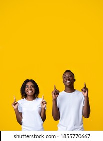Excited black couple looking up, pointing at copy space over yellow studio background. Amazed african american man and woman watching advertisement or announcement about seasonal sales and offers