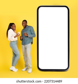 Excited Black Couple Texting On Smartphones Standing Near Huge Mobile Phone With Blank Screen Standing Over Yellow Studio Background. Great Mobile Application Advertisement. Square, Mockup