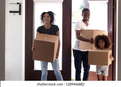 Excited black family with kid girl entering big modern house holding boxes on moving day, happy parents and child daughter standing in hallway looking around, tenants welcome to new home concept
