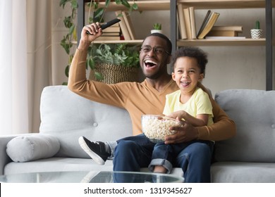 Excited Black Father With Toddler Son Fans Watching Football Sport Tv Game Celebrating Goal, Happy African Dad And Little Kid Child Cheering Favorite Soccer Team Victory At Home Sitting On Couch