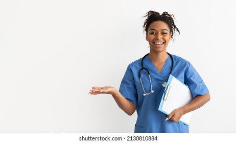 Excited black female doctor in scrubs holding clipboard showing something on empty palm pointing with hand at free copy space, advertising poduct or clinic, posing isolated on white studio background
