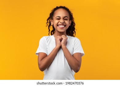 Excited Black Girl Anticipating Something Wonderful, Making Wish, Begging Or Praying Over Yellow Orange Studio Background. Hopeful African American Teen Wishing For Good Luck, Waiting For Miracle