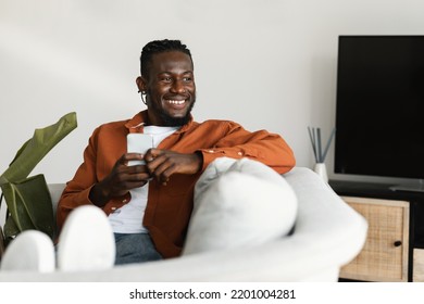 Excited Black Guy Chatting On Cellphone Or Surfing Internet, Sitting On Sofa At Home, Looking Aside At Copy Space And Smiling. Modern Technologies And Lifestyle