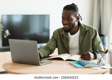 Excited Black Guy Looking For Job Online, Sitting At Table, Using Laptop Computer And Taking Notes, Copy Space. Positive African American Man Working Online From Home, Remote Job