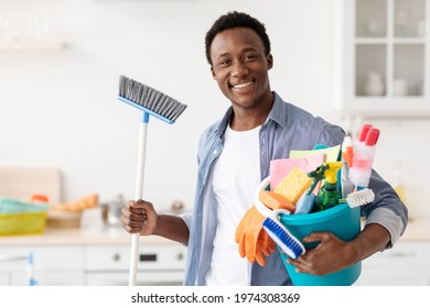 Excited black guy ready for cleaning apartment, holding mop and other cleaning tools, white cozy kitchen interior, copy space. Cheerful african american man house-keeping on weekend