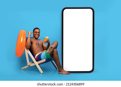 Excited black guy with tropical cocktail chilling in lounge chair near huge smartphone with blank screen for your ad, blue background. man relaxing on beach with cool drink on summer holidays