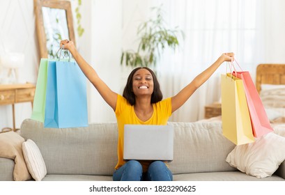 Excited Black Lady With Laptop Lifting Hands Full Of Shopping Bags On Sofa At Home. Lovely African American Woman Feeling Happy Over Big Sale In Online Shop, Purchasing Goods Over Internet Indoors