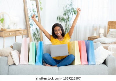 Excited Black Lady Shopping Online, Sitting On Sofa With Laptop Computer, Surrounded By Gift Bags, Indoors. Lovely African American Woman Purchasing Presents On Web, Getting Big Discount