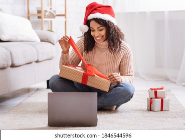 Excited Black Lady Sitting With Christmas Present And Opening It, Smiling Woman Unwrapping Gift Box With Red Ribbon In Living Room With Laptop Computer, Celebrating Online, Staying At Home