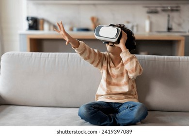 Excited black little boy wearing virtual reality headset having fun at home, african american male kid immersed in digital world sitting on sofa in bright modern living room at home, copy space - Powered by Shutterstock