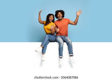 Excited black lovers sitting on horizontal white empty board for text or advertisement, happy african american man and woman screaming and gesturing, blue studio background, copy space