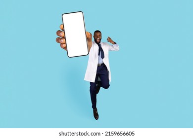 Excited Black Male Doctor Jumping With Big Blank Smartphone In Hand Over Blue Background, Cheerful African American Therapist Man In Uniform Showing White Mobile Phone Screen At Camera, Mockup