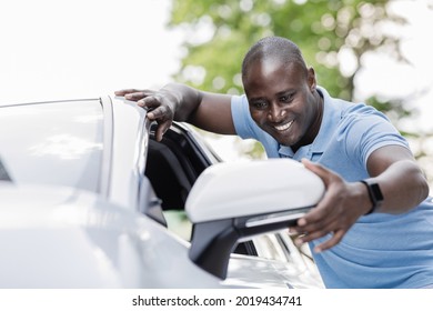 Excited black man in casual summer outfit standing outdoors by nice white car, looking at side mirror and smiling, choosing and purchasing new automobile. Car retail, auto buying concept.