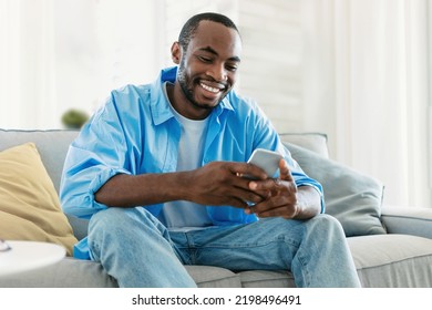 Excited Black Man Chatting On Smartphone, Watching Video, Playing Game While Resting On Sofa In Living Room Interior. Free Time For Blog At Home, New App, Advertisement And Offer