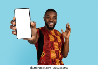 Excited Black Man Demonstrating Big Blank Smartphone In His Hand, Surprised African Guy In Traditional Costume Showing Mobile Phone With White Screen While Posing Isolated On Blue Background, Mockup