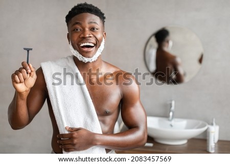 Excited Black Man With Face Covered With Shaving Foam Holding Safety Razor Removing Facial Hair Posing Smiling To Camera In Modern Bathroom At Home. Male Beauty Routine Concept