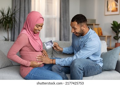 Excited Black Man Holding Baby Sonography Photo And Touching Pregnant Muslim Wife's Belly While They Relaxing Together On Couch At Home, Happy Islamic Spouses Enjoying Upcoming Parenthood, Free Space