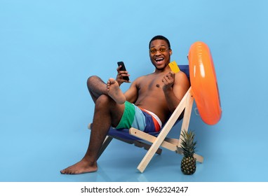 Excited Black Man Holding Credit Card And Smartphone, Shopping Online In Lounge Chair On Blue Studio Background. Cool African American Guy Buying Goods In Web Store During Summer Vacation