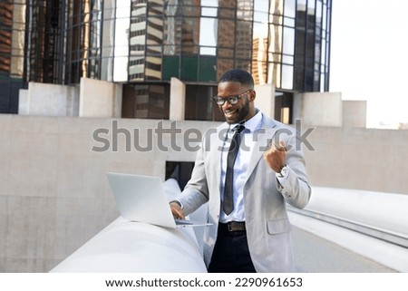 Excited Black Man Making Yes Gesture, Looking At Laptop Display, Great News. InFront OF Modern Office Buildings