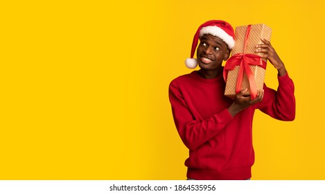 Excited Black Man In Santa Hat Shaking Wrapped Gift Box, Wonder What's Inside, Receiving Christmas Presents While Standing Over Yellow Background, Enjoying Celebrating Winter Xmas Holidays, Copy Space