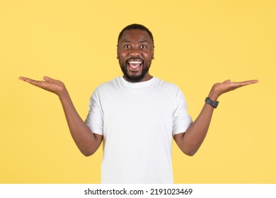 Excited Black Millennial Man Holding Hands Comparing Two Invisible Objects Or Shrugging Shoulders Smiling To Camera Posing Standing Over Yellow Background, Studio Shot