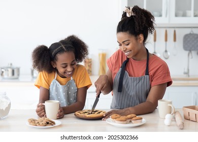 Excited black mom and daughter in aprons tasting homemade berry pie, cutting cake and smiling, kitchen interior. Happy african american mother and kid teen girl baking together at home, copy space