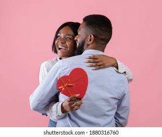 Excited black woman holding heart shaped gift box and hugging her boyfriend on pink studio background. Lovely African American couple celebrating Valentine's Day together
