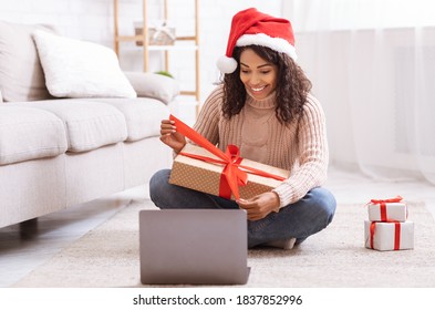 Excited Black Woman Holding And Opening Christmas Present, Smiling Lady Unwrapping Gift Box With Red Bow, Sitting On The Floor At Home In Living Room With Laptop Computer, Celebrating Online