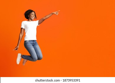 Excited black woman jumping up and showing copy space over orange studio background. Emotional african american millennial lady smiling and pointing at advertising or announcement