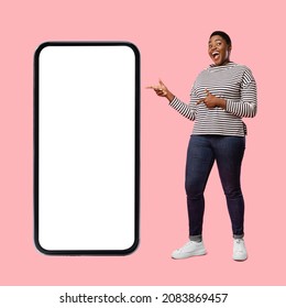 Excited Black Woman Near Big Smartphone Pointing Fingers At Huge Blank Phone Screen Advertising Mobile Application Standing Over Pink Background. Studio Shot, Square