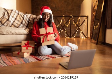 Excited Black Woman Opening Christmas Gifts In Front Of Laptop, Celebrating Xmas Or New Year Online At Home, Copy Space. Positive Young Lady Unwrapping Presents While Video Chatting On Web