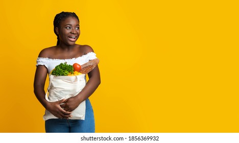 Excited Black Woman With Paper Bag Of Groceries In Hands Looking Away At Copy Space, Enjoying Market Discount Sale Offer For Food Shopping, Standing Over Yellow Studio Background, Panorama