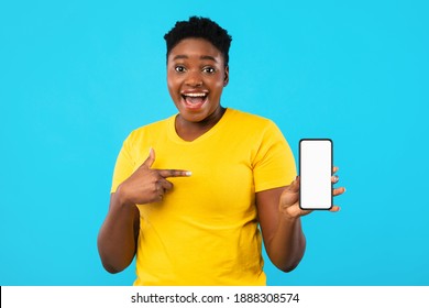 Excited Black Woman Pointing At Phone Blank Screen Showing Smartphone To Camera Recommending Application Over Blue Studio Background. Great Mobile App For Cellphone Concept. Mockup