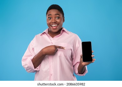 Excited Black Woman Showing Phone Blank Screen, Pointing Finger Recommending Mobile Application Standing On Blue Background. Check This App For Smartphone Concept. Studio Shot, Mockup