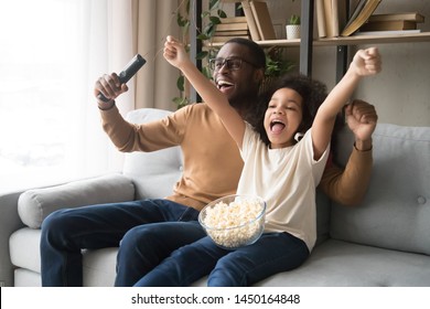 Excited Black Young Dad And Preschooler Daughter Eat Popcorn Watching Football Match Together, Happy African American Father And Girl Child Relax On Couch Cheering Team Enjoy Game On TV At Home
