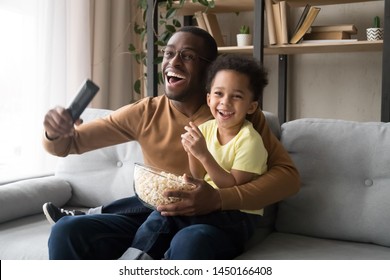 Excited Black Young Father And Preschooler Son Sit On Couch Eating Popcorn Enjoy Football Match On TV Cheering, Smiling Overjoyed African American Dad And Funny Boy Child Fans Watch Game At Home