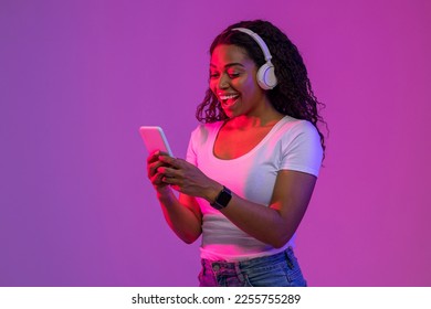 Excited Black Young Woman Wearing Wireless headphones Using Smartphone In Neon Light, Happy Surprised African American Female Browsing New Application Or Messaging With Friends, Copy Space - Powered by Shutterstock