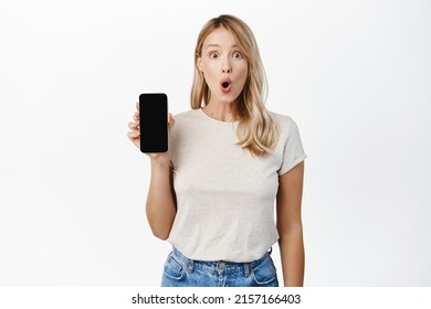 Excited Blond Girl Showing Mobile Phone Screen, Say Wow, Demonstrating Smartphone App Interface, Standing Over White Background