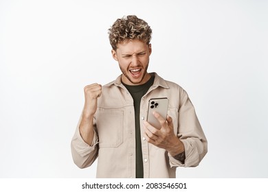 Excited blond man looking at mobile phone and triumphing, watching video and winning bet, makng fist pump, achieve goal on smartphone, standing over white background