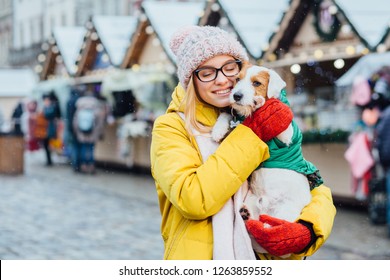 Excited Blond Sweden Woman In Warm Yellow Jacket, Red Mittens, Knitted Hat Holding And Hugging Her Adorable Jack Russel Terrier Dog In Green Outwear During Winter Snowy Day Over Christmas Market
